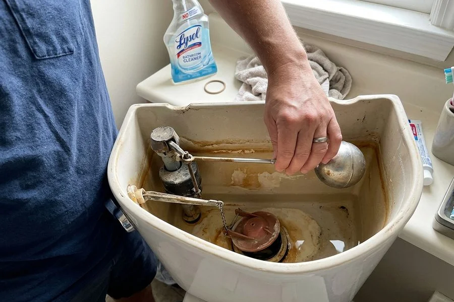 Open toilet tank showing a worn flapper and fill valve being adjusted
